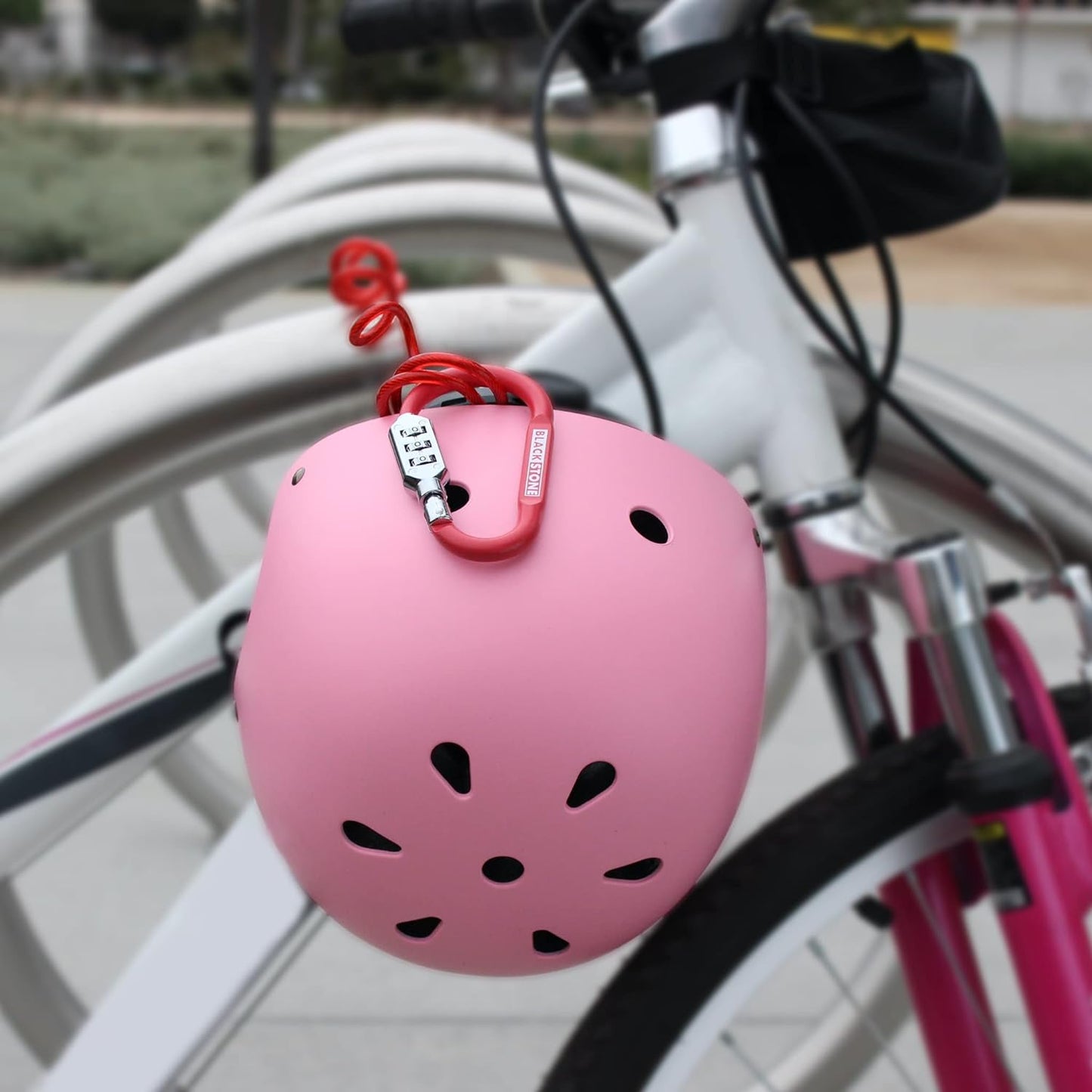 Pink helmet secured with a red Black Stone combination lock to a white bike on a bike rack, with soft focus on the surrounding environment