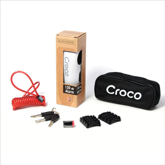 The image shows a product set which includes a Blackstone Croco alarm lock in its packaging, a red coiled cable, a black carrying pouch with the text "Croco" on it, three keys, a battery, and Two rubber pads for the lock, offering customization for various handlebar thicknesses or preferences. The items are neatly displayed, presumably for marketing or sales purposes