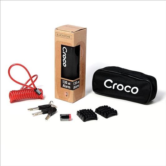 The image shows a product set which includes a Blackstone Croco alarm lock in its packaging, a red coiled cable, a black carrying pouch with the text "Croco" on it, three keys, a battery, and Two rubber pads for the lock, offering customization for various handlebar thicknesses or preferences. The items are neatly displayed, presumably for marketing or sales purposes