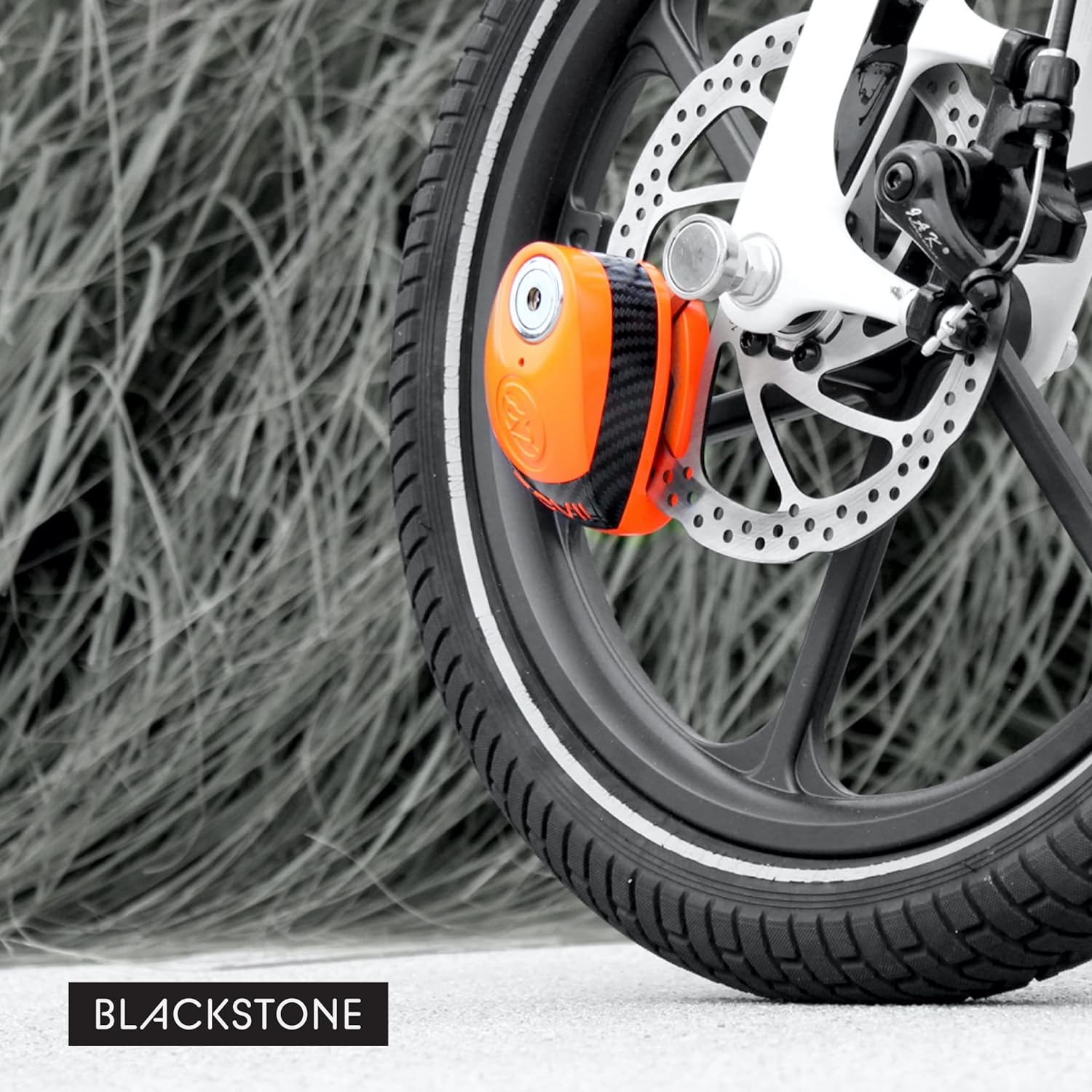 In this image, we see a modern and the bold red disc lock attached to the front brake disc of a motorcycle. The color of the lock provides a striking contrast against the grayscale background, drawing the viewer’s attention immediately to the security device. The disc lock is compact, yet its placement suggests a robust and effective deterrent against theft. The "BLACKSTONE" branding is discreetly positioned in the lower right corner, suggesting a focus on the product's sleek design and functionality
