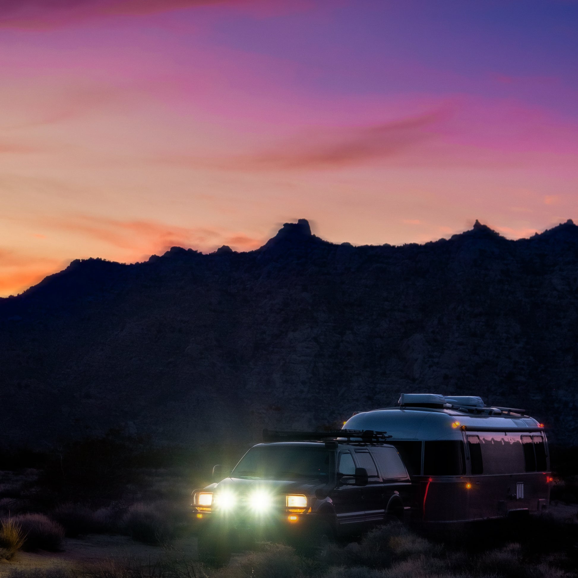 An RV camping in the wilderness at dusk with the mountains in the background under a purple sky. The vehicle's headlights are on, creating a sense of adventure and solitude in the vast outdoors